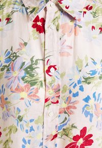 Chemise claire boutonnée avec un motif floral multicolore comprenant des fleurs et des feuilles rouges, bleues, roses et vertes.