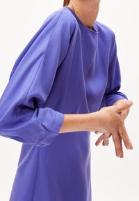 Abito viola con scollo a giro e maniche a sbuffo. Il tessuto ha una texture liscia e un sottile motivo a righe. Dettaglio con bottoni sui polsini.