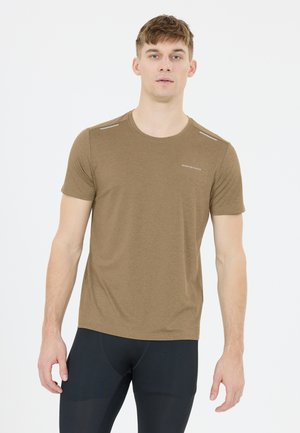Mann steht vor weißem Hintergrund, trägt ein braunes kurzärmliges Sportshirt mit dem Text "Endurance" und schwarze enganliegende Hose.