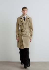 Béžový trenčkot s dvojradovým zapínaním na gombíky a opaskom v páse. Má široké klopy a hladký materiál, v kombinácii s čiernymi nohavicami.