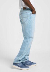 Jeans de denim azul claro con un diseño de pierna recta, que cuentan con bolsillos traseros y un parche de cuero marrón en la cintura. Llevados con zapatillas deportivas azules.