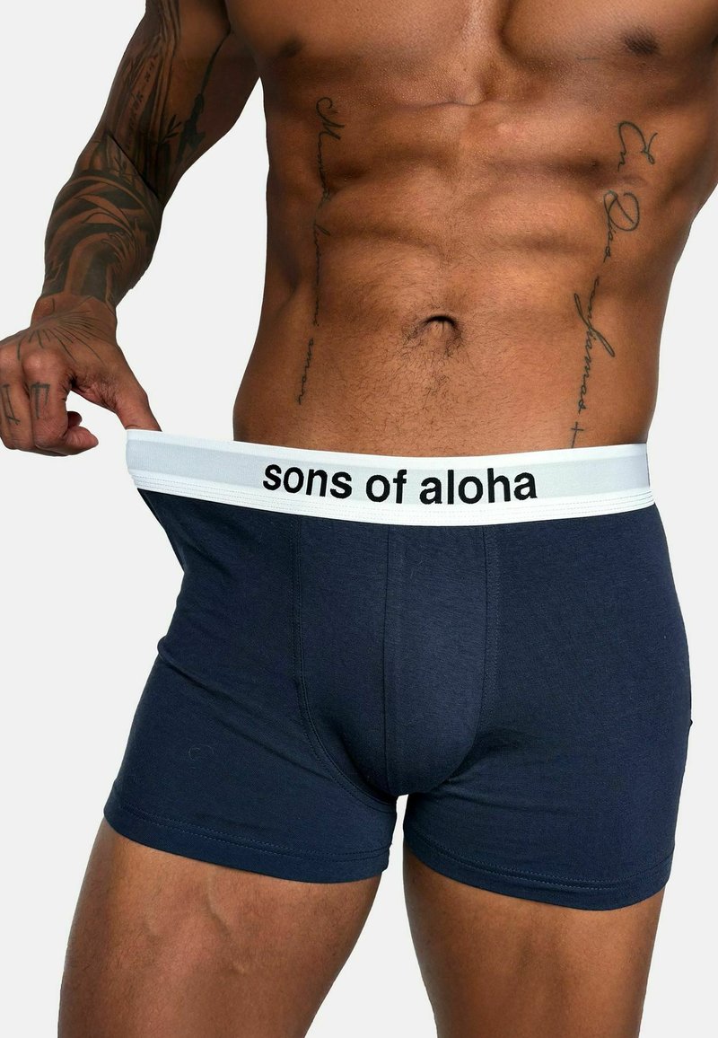 Boxer shorts blu navy con una fascia elastica bianca riportante la scritta "sons of aloha". Il tessuto appare liscio e aderente.
