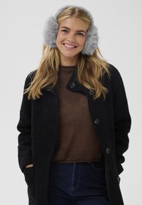 Cappotto nero con trama e grandi bottoni, maglione marrone lavorato a maglia e paraorecchie grigi in pelliccia sintetica indossati da una donna sorridente. Jeans blu visibili.