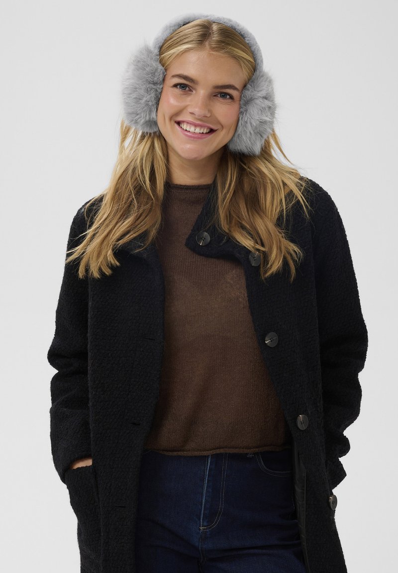 Cappotto nero con trama e grandi bottoni, maglione marrone lavorato a maglia e paraorecchie grigi in pelliccia sintetica indossati da una donna sorridente. Jeans blu visibili.