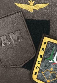 Toppa in pelle marrone con la stampa in rilievo delle lettere "AM", emblema giallo ricamato di un'aquila e un distintivo colorato "Frecce" con accenti verdi, bianchi e rossi.