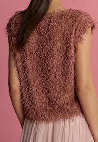 Top senza maniche color rosa polveroso con dettagli in frangia indossato con una gonna a pieghe rosa chiaro, mostrato di schiena su uno sfondo rosa.