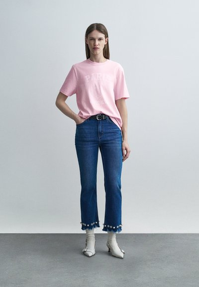 Γυναίκα που στέκεται φορώντας ένα ροζ T-shirt με την επιγραφή «Paris», μπλε ψηλόμεσα τζιν με λεπτομέρειες από πέρλες, λευκές κάλτσες και λευκά ψηλοτάκουνα παπούτσια.