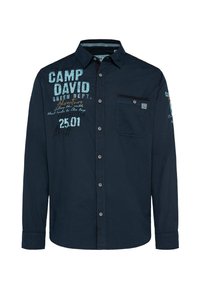 Maglietta a maniche lunghe navy con taschino frontale, chiusura a bottone e testo grafico in blu chiaro e arancione. Tessuto di cotone morbido e informale.