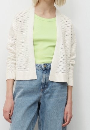 Leichter, gestrickter Cardigan in Creme, mit offener Vorderseite und gerippten Bündchen. Über einem hellgrünen Oberteil getragen und mit blauen Jeans kombiniert.