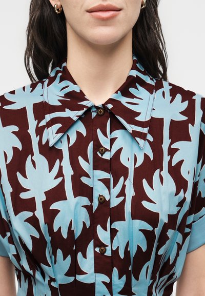 Γυναίκα που φοράει πουκάμισο με κουμπιά και μεγάλο γιακά με μπλε σχέδια φοίνικα σε σκούρο μαροκινό φόντο.