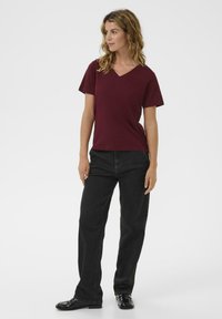Burgundy v-neck t-shirt made of cotton, paired with high-waisted dark denim jeans. Model stands on a plain background, wearing black shoes.