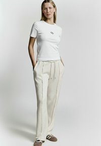 Weißes, tailliertes T-Shirt mit kurzen Ärmeln und schwarzem Logo. Cremefarbene Jogginghose mit seitlichen Nähten, Kordelzugbund und Taschen.