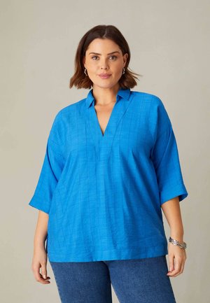 Blue short-sleeve top with a V-neck and textured fabric, featuring a relaxed fit and vertical striations, paired with blue jeans.