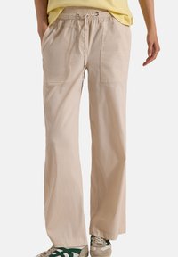 Personne portant un pantalon beige à cordon avec de grandes poches à l'avant, une chemise jaune et des baskets beiges à bandes vertes, les mains dans les poches.