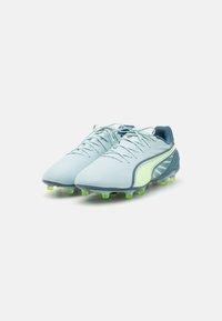Scarpe sportive con una parte superiore di un blu chiaro, dettagli verdi e una suola texturizzata con molteplici tacchetti per garantire aderenza su superfici erbose.