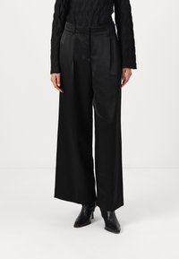 Pantaloni neri a gamba larga realizzati in un tessuto liscio e lucido, con una vita su misura e pieghe frontali, abbinati a stivaletti neri.