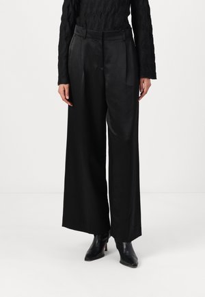 Pantalons larges noirs en tissu lisse et brillant, dotés d'une taille ajustée et de plis à l'avant, associés à des bottines noires.