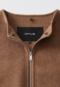 Braune Wolljacke mit partiell geöffnetem silbernen Reißverschluss, Knopfdetails am Kragen und schwarzem Etikett mit der Aufschrift "OPUS" und Größenetikett "S".