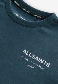 Tamsiai mėlynas medvilninis marškinėliai su apvaliu kaklu. Priekyje yra baltas užrašas "ALLSAINTS" ir papildomas tekstas japonų kalba, esantis centre.
