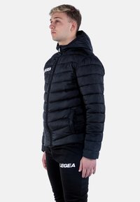 Svart puffjacka med huva, med dragkedjestängning och horisontell quiltad mönster. Visas på en modell som bär matchande svarta byxor.