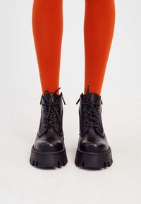 Black lace-up ankle boots with a chunky, ridged sole and rope detailing, set against bright orange tights. Smooth leather upper, minimalistic design.