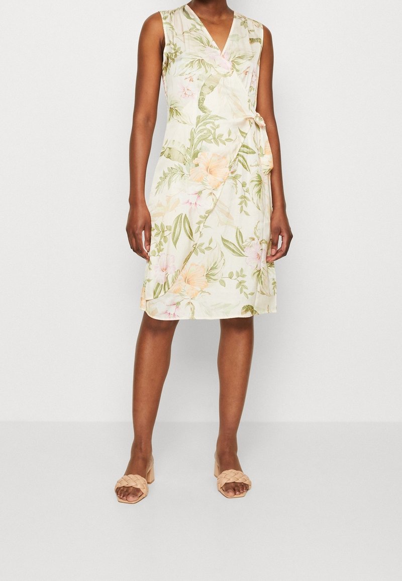 Femme portant une robe portefeuille sans manches, longueur genoux, à motif floral aux tons verts et pêche, associée à des sandales beige à talons blocs avec bride tressée.