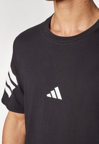 Černé bavlněné tričko s kulatým výstřihem, bílým trojúhelníkovým logem na přední straně a třemi bílými pruhy na levém rukávu.