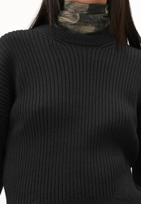 Pull noir côtelé avec un col large, associé à un col roulé camouflage vert. Le tissu semble doux avec une finition texturée.