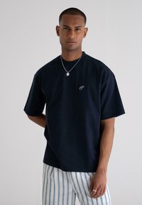 Navy-blue T-shirt with short sleeves and relaxed fit. Features a small white logo on the chest. Paired with cream and blue striped pants.