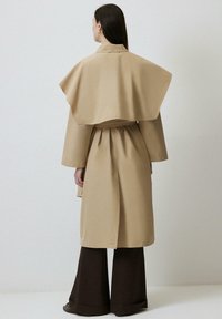 Palton bej cu un guler unic de tip capa și talie accentuată. Mâneci lungi și o croială elegantă, asortat cu pantaloni largi de culoare maro închis.