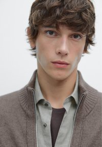 Brauner Cardigan mit Reißverschluss über einem hellgrünen Hemd und einem dunklen T-Shirt darunter. Der Stoff wirkt weich, mit einem strukturierten, gerippten Kragen.