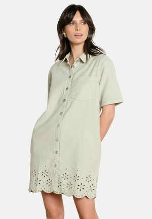 Woman wearing a light beige button-up shirt dress with short sleeves and floral cutout patterns along the scalloped hem.