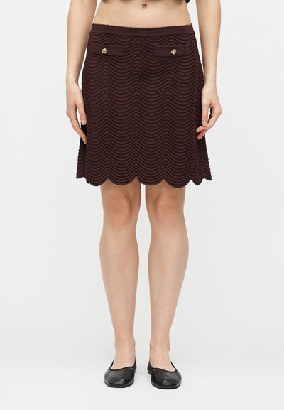 Jupe brun foncé texturée ondulée longueur genoux avec ourlet festonné, deux poches avant avec boutons dorés, portée avec des ballerines noires.