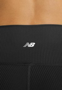 Primer plano de la parte baja de la espalda de una persona que lleva leggings deportivos negros de canalé, con un pequeño logo blanco "NB" en el centro de la cinturilla.