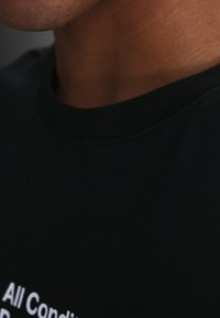 Κοντινό πλάνο ενός ατόμου που φοράει μαύρο πουκάμισο με μερικώς ορατό λευκό κείμενο κοντά στον γιακά και τον λαιμό.