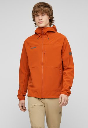 Junger Mann trägt eine orange Kapuzenjacke mit Reißverschlusstaschen und beigen Hosen vor einem schlichten hellgrauen Hintergrund.
