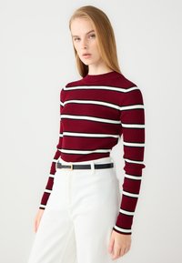 Maglia a maniche lunghe rossa a coste con righe orizzontali bianche e nere, abbinata a pantaloni bianchi a vita alta e una cintura nera.