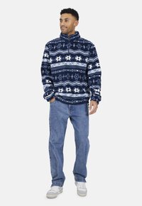 Pullover in pile blu navy con motivo geometrico bianco, colletto con zip a metà e polsini a coste, abbinato a jeans in denim blu chiaro e sneakers.