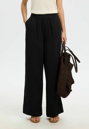 Osoba nosząca czarne spodnie o szerokich nogawkach z wysokim stanem i beżowy top, trzymająca dużą ciemną plecioną torbę na ramię jedną ręką.