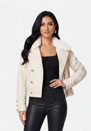 Vrouw met lang zwart haar draagt een crème cropped jas met witte nepbontkraag, gouden knopen, zwarte top en zwarte leren broek.