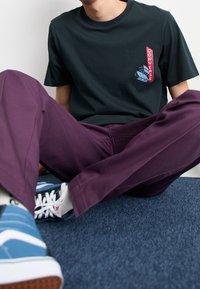 Svart bomullst-shirt med röd "Converse"-grafik och blå sneakersdetaljer, kombinerad med lila byxor, sittande på blå texturerad yta.