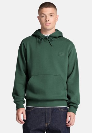 Joven con una sudadera con capucha de color verde oscuro con un bolsillo frontal y vaqueros azul oscuro frente a un fondo claro y liso.