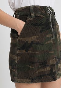 Jupe en velours côtelé à motif camouflage en vert et marron, dotée d'une fermeture éclair à l'avant et de poches latérales ; tissu texturé avec une silhouette ajustée.
