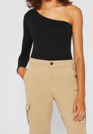 Vrouw draagt een zwarte top met één blote schouder en lange mouwen en beige cargobroek met handen in de zakken tegen een effen achtergrond.