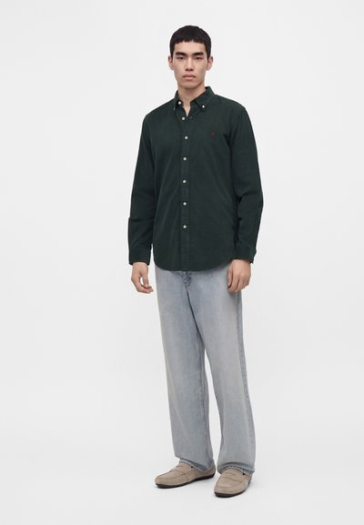 Jeune homme debout portant une chemise boutonnée vert foncé, un jean large bleu clair et des chaussures beiges sans lacets sur un fond blanc uni.