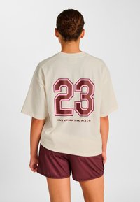 Cremefarvet T-shirt med korte ærmer med stort mørkerødt "23" og "INTERNATIONALS" trykt på ryggen, kombineret med mørkerøde shorts.