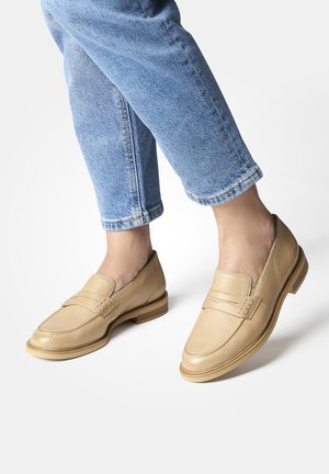 Mocassini in pelle beige con finitura liscia, caratterizzati da un design cucito e un tacco in legno, abbinati a jeans in denim blu arrotolati.