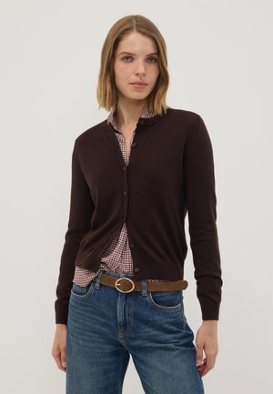 Frau trägt eine dunkelbraune Strickjacke über einem rot-weißen karierten Hemd, blaue Jeans und einen braunen Gürtel mit einer runden goldenen Schnalle.