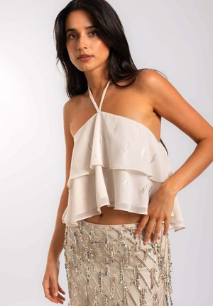 Top sin mangas de color beige claro y con capas, con cuello halter; presenta acentos plateados y una silueta fluida. Combinado con una falda de cuentas.