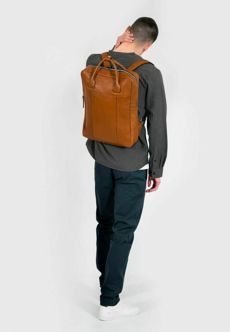 Sac à dos en cuir marron avec deux bretelles, une poignée supérieure et une forme rectangulaire. Présente une texture lisse et des lignes épurées.